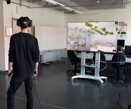 Eine Person hat eine VR-Brille auf und steht in einem Raum. Auf einem Bildschirm im Hintergrund ist die VR-Umgebung sichtbar.